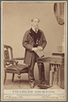 Full-length portrait of Charles Dickens, standing at desk, clasping book in both hands