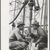 Oil drillers talking with bits in front of them and drilling equipment in background, Kilgore, Texas