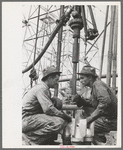 Oil drillers talking with bits in front of them and drilling equipment in background, Kilgore, Texas