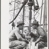 Oil drillers talking with bits in front of them and drilling equipment in background, Kilgore, Texas