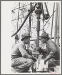Oil drillers talking with bits in front of them and drilling equipment in background, Kilgore, Texas
