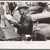 Man sitting on automobile fender reading a newspaper. Market square, Waco, Texas