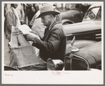 Man sitting on automobile fender reading a newspaper. Market square, Waco, Texas