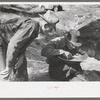 Gold prospector blowing away dirt to find the gold in his pan while a visiting prospector looks on, Pinos Altos, New Mexico