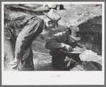 Gold prospector blowing away dirt to find the gold in his pan while a visiting prospector looks on, Pinos Altos, New Mexico