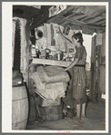 Woman living in shack at Tin Town, Caruthersville, Missouri, in front of shelf used as kitchen cabinet. Note ceiling and dirt floor