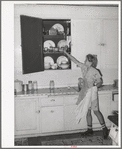 Daughter of Mormon farmer putting away dishes in kitchen cabinet. Box Elder County, Utah