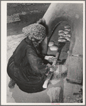 Spanish-American woman removing baked bread from outdoor earthen oven by means of a long wooden paddle. Near Taos, New Mexico