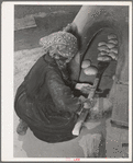 Spanish-American woman removing baked bread from outdoor earthen oven by means of a long wooden paddle. Near Taos, New Mexico