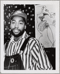 Half-length portrait of fashion designer Patrick Kelly, wearing a striped shirt, overalls, and Nike baseball cap, and standing in front of a large drawing, during a visit at Howard University where he spoke to fashion students