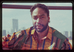 Portrait of fashion designer Patrick Kelly, wearing kente print button up shirt, with city skyline in background