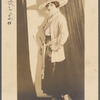 Julian Eltinge in broad-brimmed hat wearing a loose cardigan, dark skirt and heels, carrying a rod or parasol with curtain backdrop
