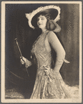 Julian Eltinge in brocaded dress with arm-coverings wearing broad-brimmed hat with feather frills and holding baton
