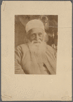 Photograph of man with beard in brimless hat and cloak, seated and facing camera
