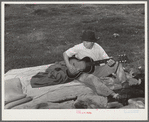 Migrant boy playing the guitar on pallet while camped near Prague, Oklahoma. Lincoln County, Oklahoma