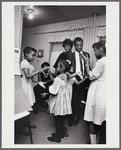 Baldwin standing in kitchen with extended family, with two pairs of girls playing hand-clapping games