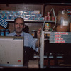 Grocer behind register with Greek flag in background, Grocer #3