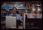 Grocer behind register with Greek flag in background, Grocer #3