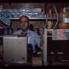 Grocer behind register with Greek flag in background, Grocer #3