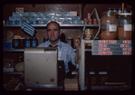 Grocer behind register with Greek flag in background, Grocer #3