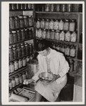 Mrs. Richard R. Gill, Donley County, Texas, prepares eggs for the market. The large containers are filled with homemade lard for this work grant family. Note that Mrs. Gill's attractive jacket is made from a sugar sack.