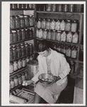 Mrs. Richard R. Gill, Donley County, Texas, prepares eggs for the market. The large containers are filled with homemade lard for this work grant family. Note that Mrs. Gill's attractive jacket is made from a sugar sack.