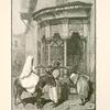 A street drinking-fountain, Damascus.  The lady who is riding en cavalier wears a dark muslin veil, called a mandîl, which quite conceals her features; her outer garment, an izzar, is like a large sheet of fine white calico, and in this she envelopes herself completely.