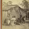 Family group sitting in front of a wooden shack.
