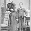 Unidentified standing figures: woman, her folded arms leaning on back of chair, and young man, probably her son, left hand on book.]