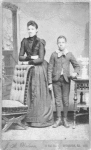 Unidentified standing figures: woman, her folded arms leaning on back of chair, and young man, probably her son, left hand on book.
