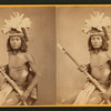 Studio portrait of Hesh-pid-die with feather headress and holding a rifle.