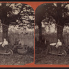A child sitting in a wheelbarrow with her doll, ax sharpener, rake, basket and watering can nearby.