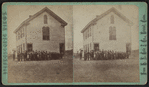 View of a two-story wood-frame school house with students and teachers out front.