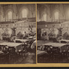 Interior of a large hall showing booths set up for a fair. (Dining tables in foreground).