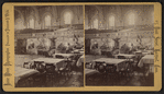 Interior of a large hall showing booths set up for a fair. (Dining tables in foreground).