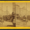 Street scene with wagons, buildings. A sign advertising liquor,cigars and tobacco in foreground.