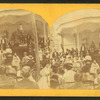 Crowd facing a bandstand where speakers and a woman at piano sit.