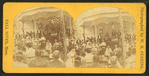 Crowd facing a bandstand where speakers and a woman at piano sit.
