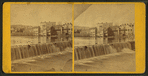View of a waterfall with buildings lining the river on the opposite shore.