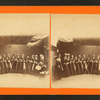 Studio view of a group of women holding brooms(?) and 1 woman with a drum.