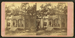 View of a cottage, trees in front, people on porch and balcony.
