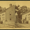 Group posing on porch of large house, possibly a boarding house, with wagonette standing by.]
