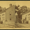 Group posing on porch of large house, possibly a boarding house, with wagonette standing by.