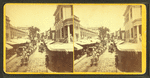 View of a commercial street lined with awmings, large horse team and wagon.
