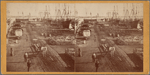 Barrels and cargo on piers, ships in background.