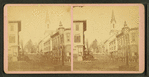 View of people standing on a commercial street, church visible in background.