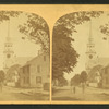View of people standing on a commercial street, church visible in background.