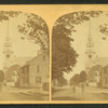 View of people standing on a commercial street, church visible in background.