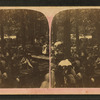 View of people standing and sitting at long tables under trees.