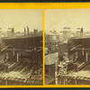 View of a house under construction, showing men finishing the chimney and building a brick wall.